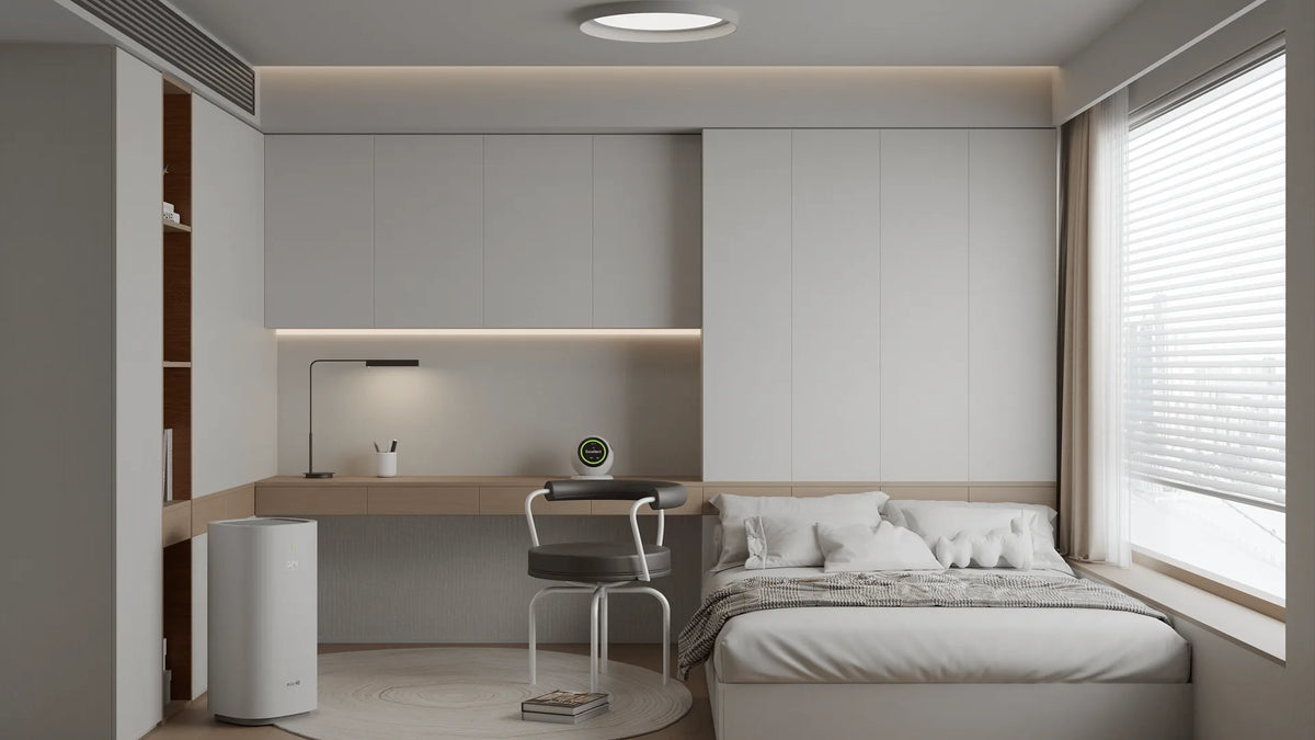 Compact room with pale built-in joinery, an oak desk ledge and task lamp running to a low bed, and a curtained window with venetian blinds, a white air purifier standing on the round rug and a white spherical air monitor showing a green Excellent reading on the desk.