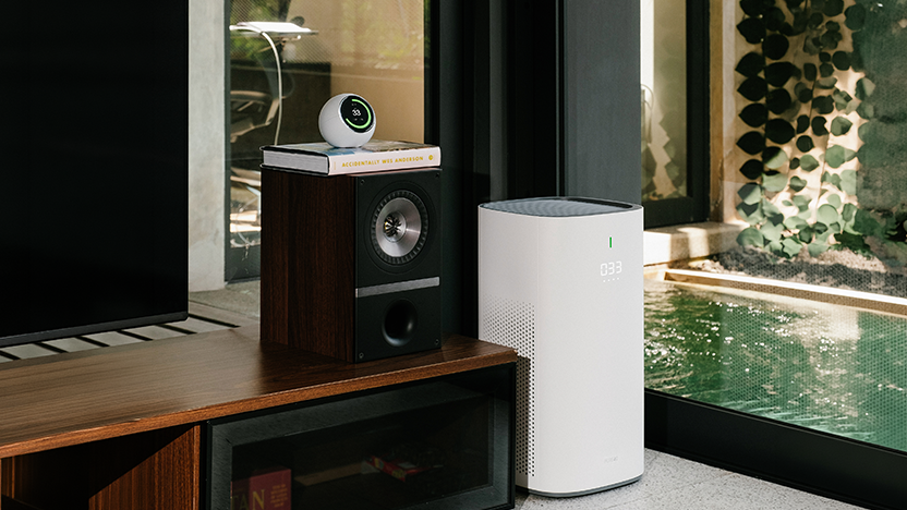 Living room with a white floor-standing air purifier reading 033 beside a garden window, and a white palm-sized spherical air monitor reading 33 in a green ring resting on a speaker cabinet beside the television.