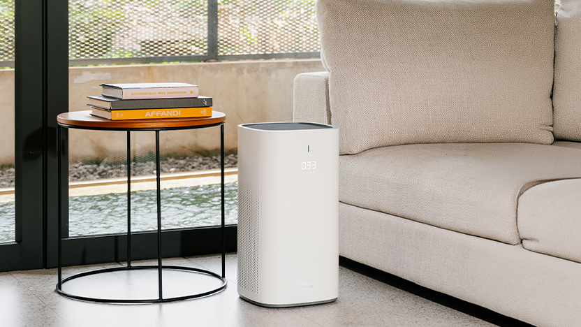 Living room with a slim white floor-standing air purifier reading 033 standing between a round side table stacked with books and a linen sofa, next to a garden window.